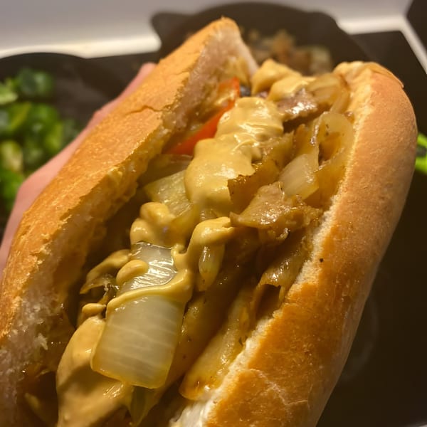 A hand holds a vegan cheesesteak sandwich with melted cheese, onions, and peppers.