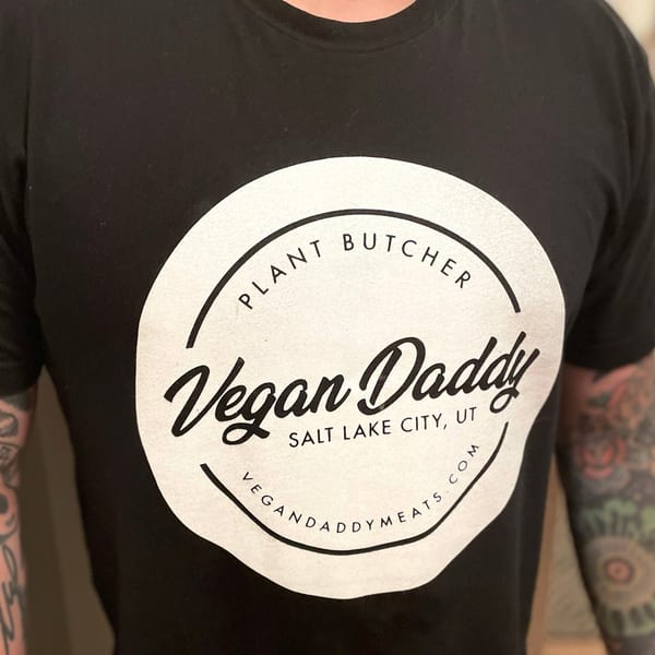 Black t-shirt with a white circular Vegan Daddy logo, worn by a person with tattooed arms.