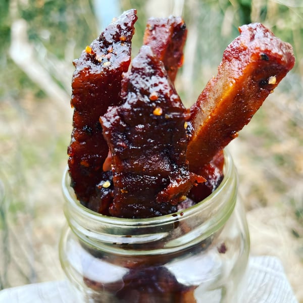 Vegan Daddy Meats' vegan bacon strips, glossy with sauce and spices, in a glass jar outdoors.
